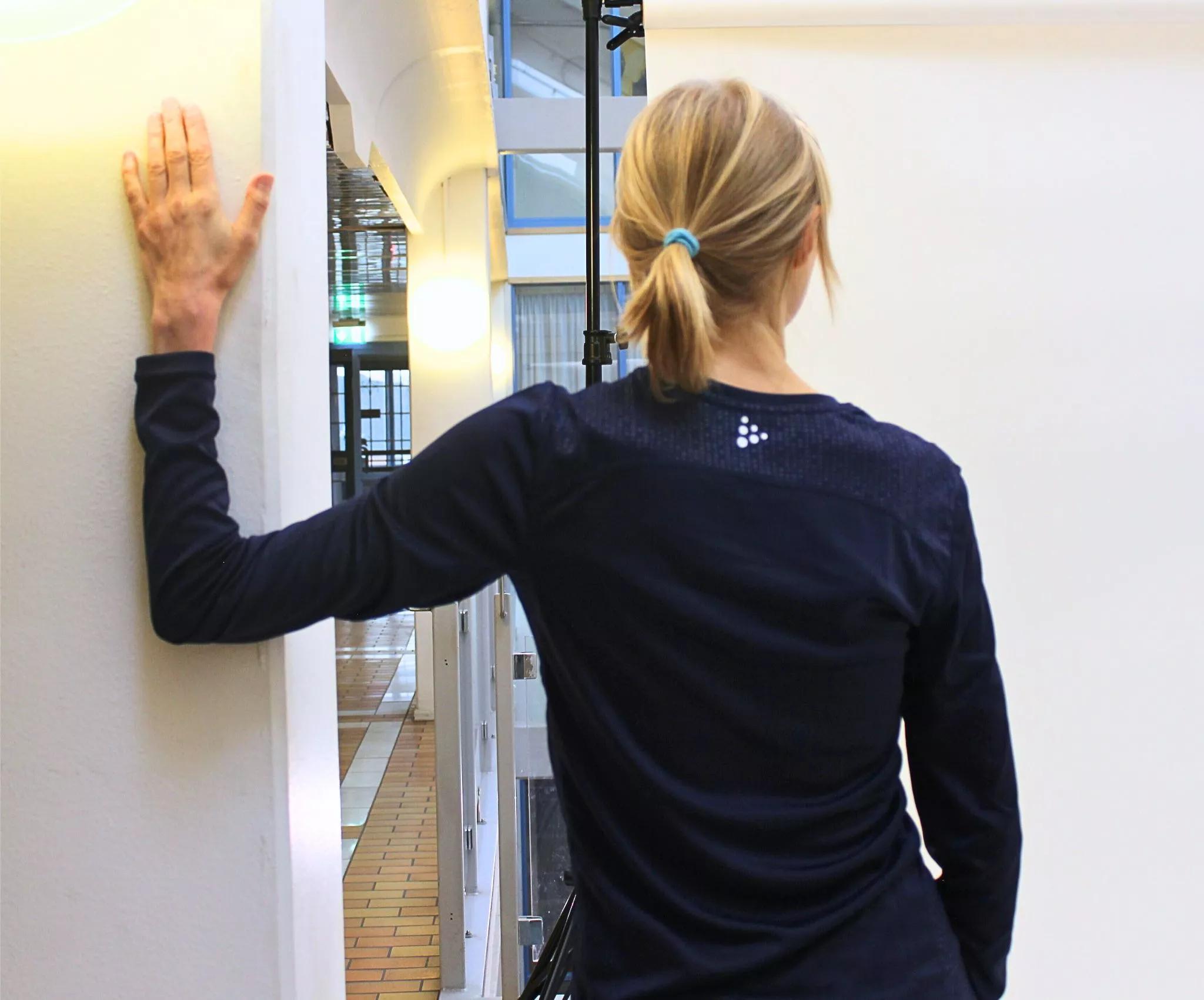 Frau zeigt Dehnung der Brustmuskeln mit Hand gegen Wand